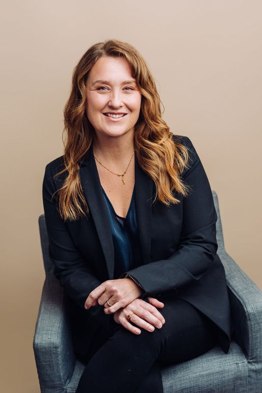 A woman in a suit is sitting in a chair with her legs crossed and smiling.