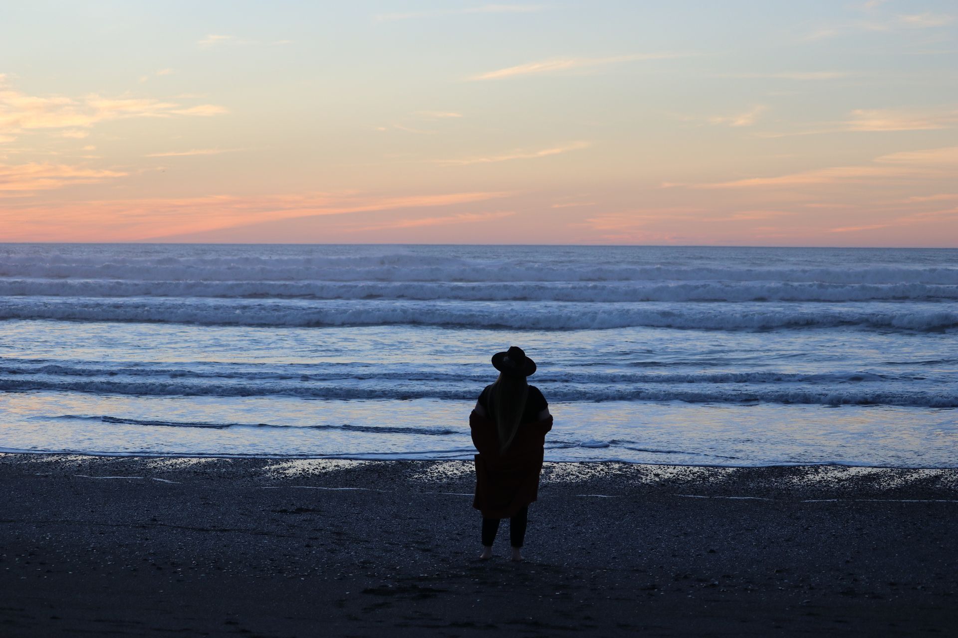 A person is standing on a beach looking at the ocean at sunset.