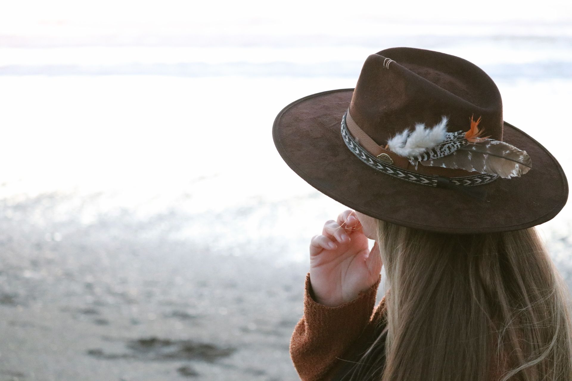 A woman wearing a brown hat with feathers on it is standing on a beach.