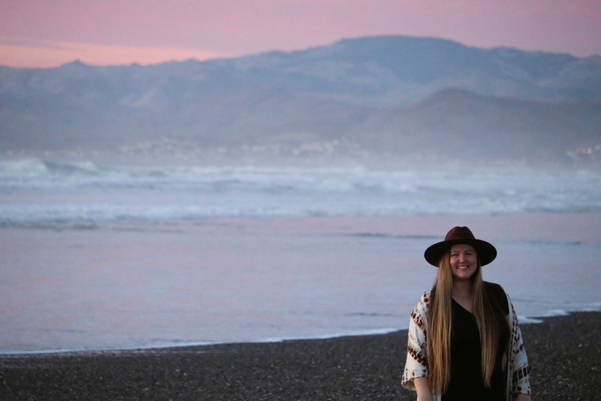 A woman in a hat is standing on a beach at sunset.