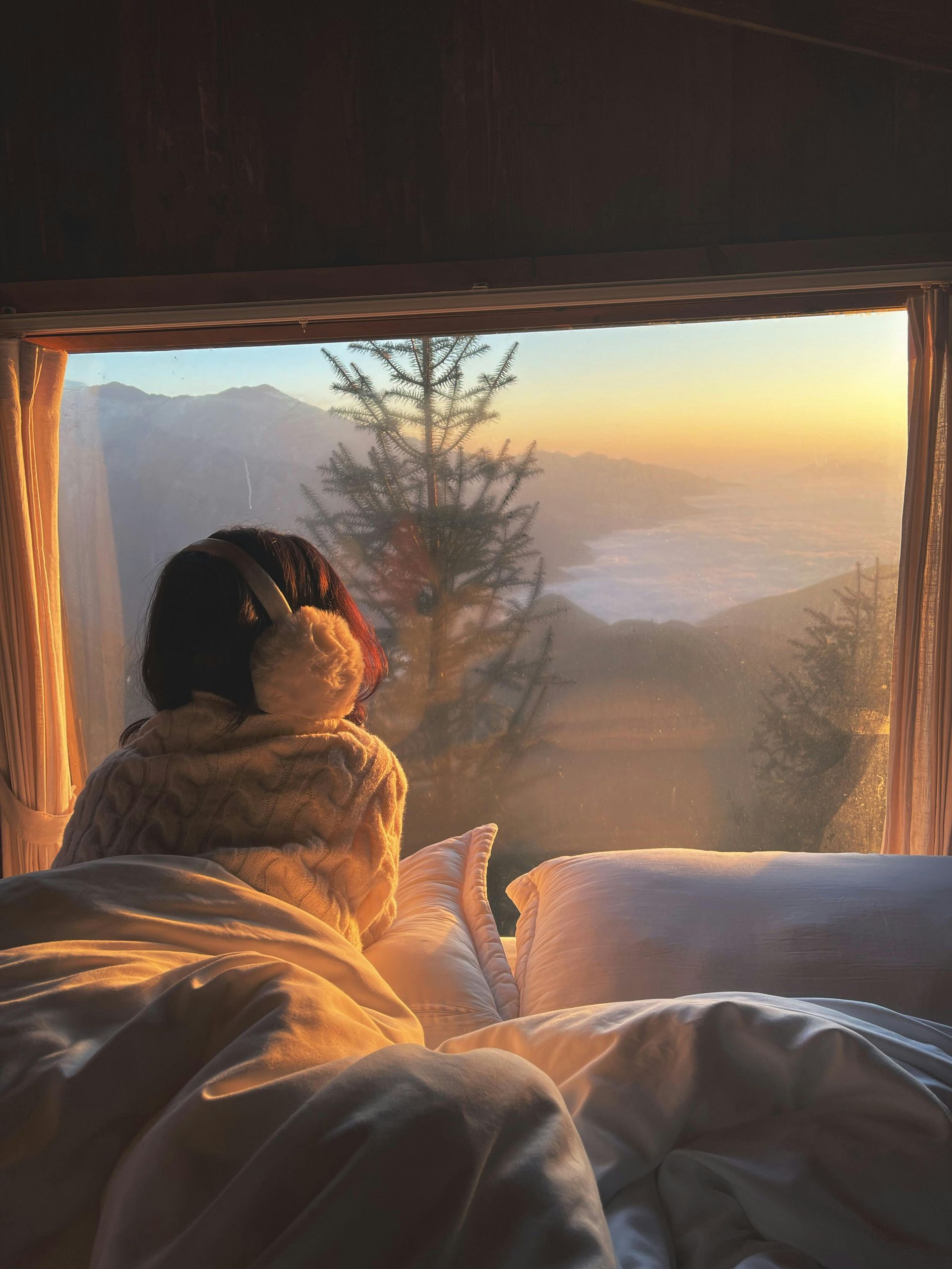 A person is laying in bed looking out of a window at a sunset.
