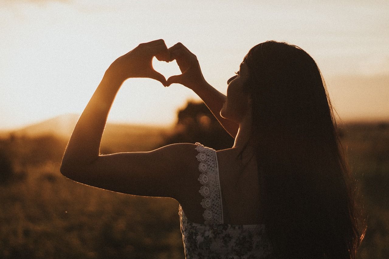 A woman is making a heart shape with her hands at sunset.