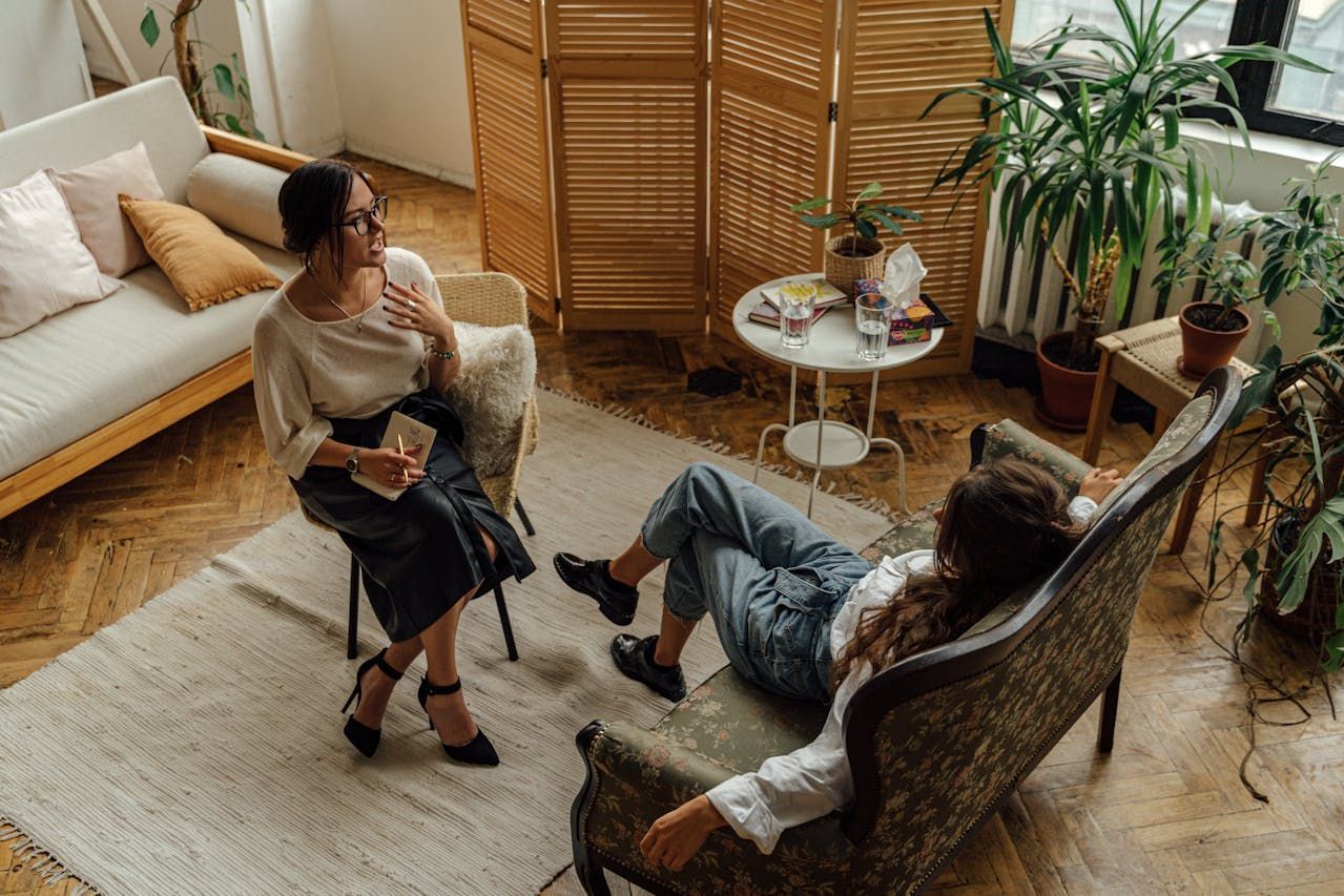 A woman is sitting in a chair talking to another woman who is laying on a couch.