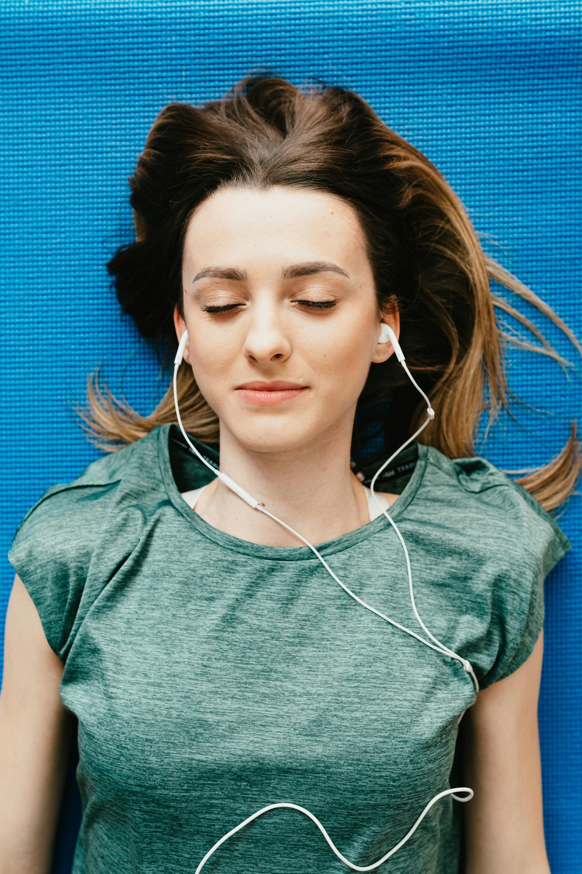 A woman is laying on a yoga mat listening to music with her eyes closed.