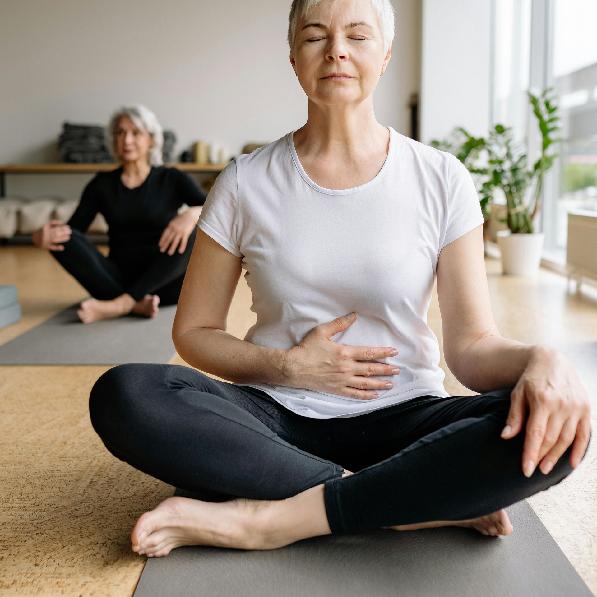 Two women are sitting on yoga mats with their eyes closed.