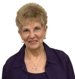 An older woman wearing a purple jacket and necklace smiles for the camera