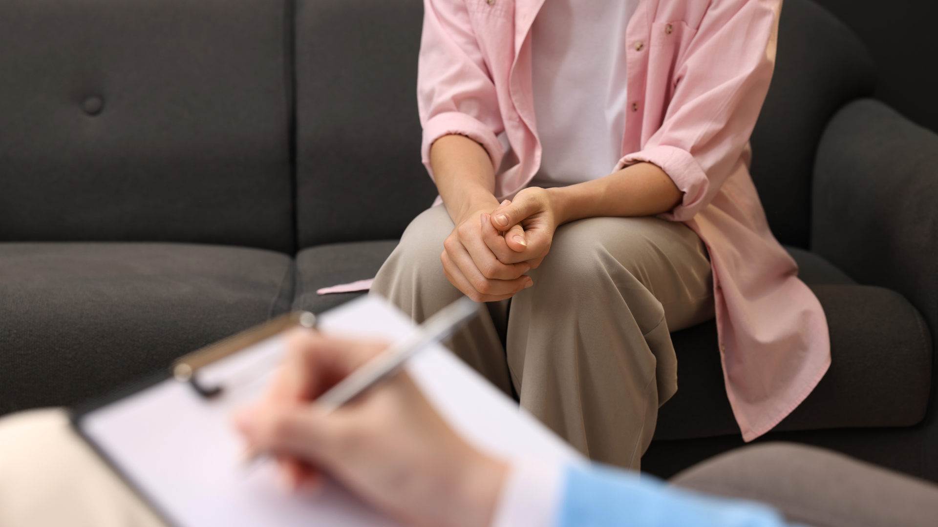 Person seated on a gray couch during a counseling session, with a blurred therapist holding a clipboard in foreground
