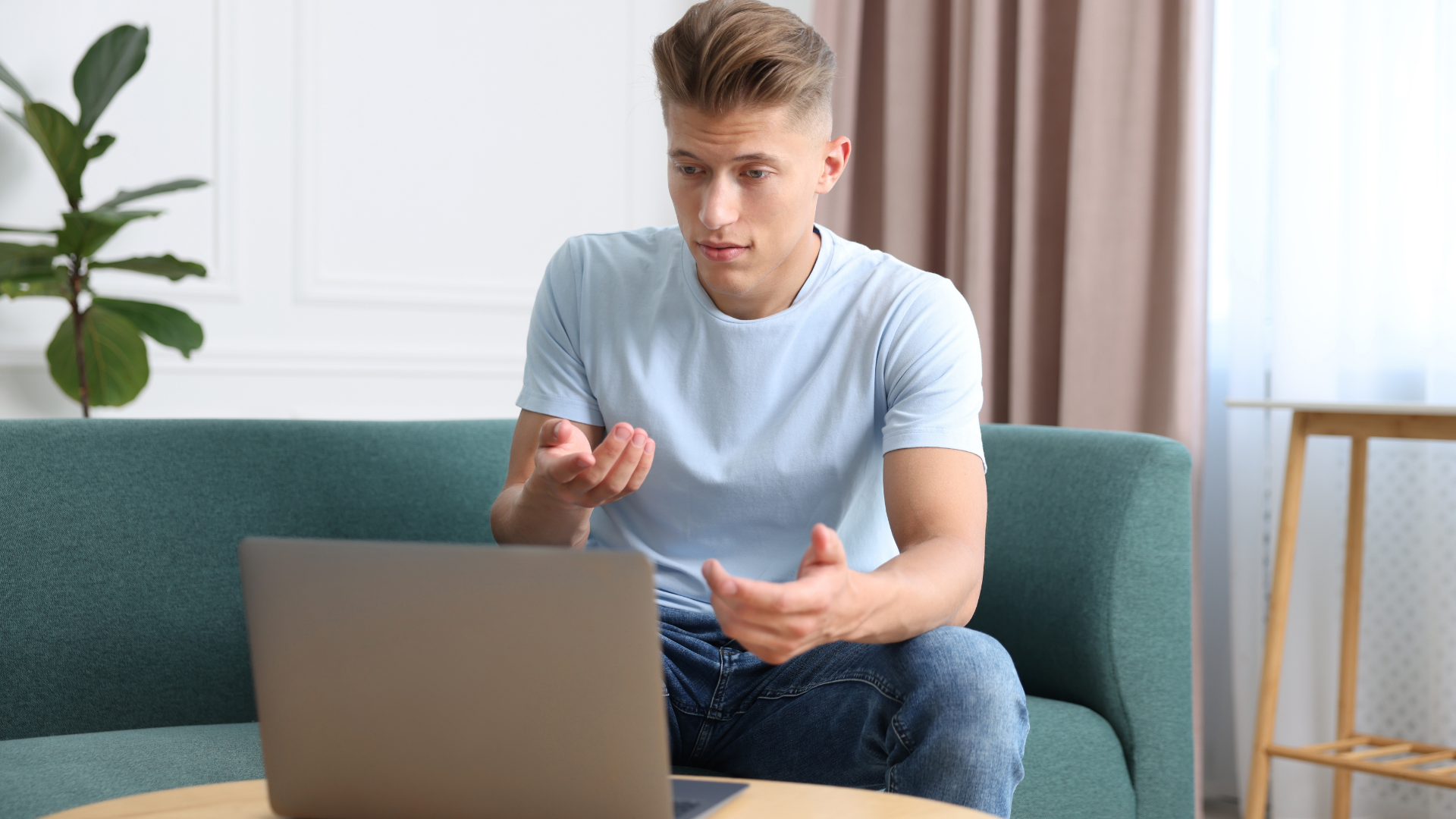 Man sitting on a sofa using a laptop, gesturing while talking in a bright living room.