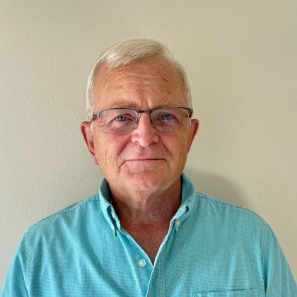 A man wearing glasses and a blue shirt is standing in front of a white wall.