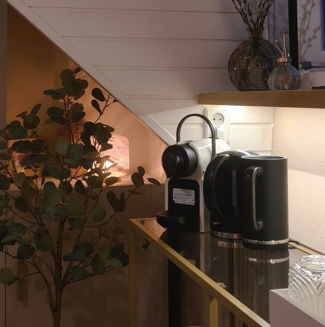 Knus hoekje in de keuken met een koffiezetapparaat en hanglampen onder een schuin plafond, naast een plant met weelderige bladeren.