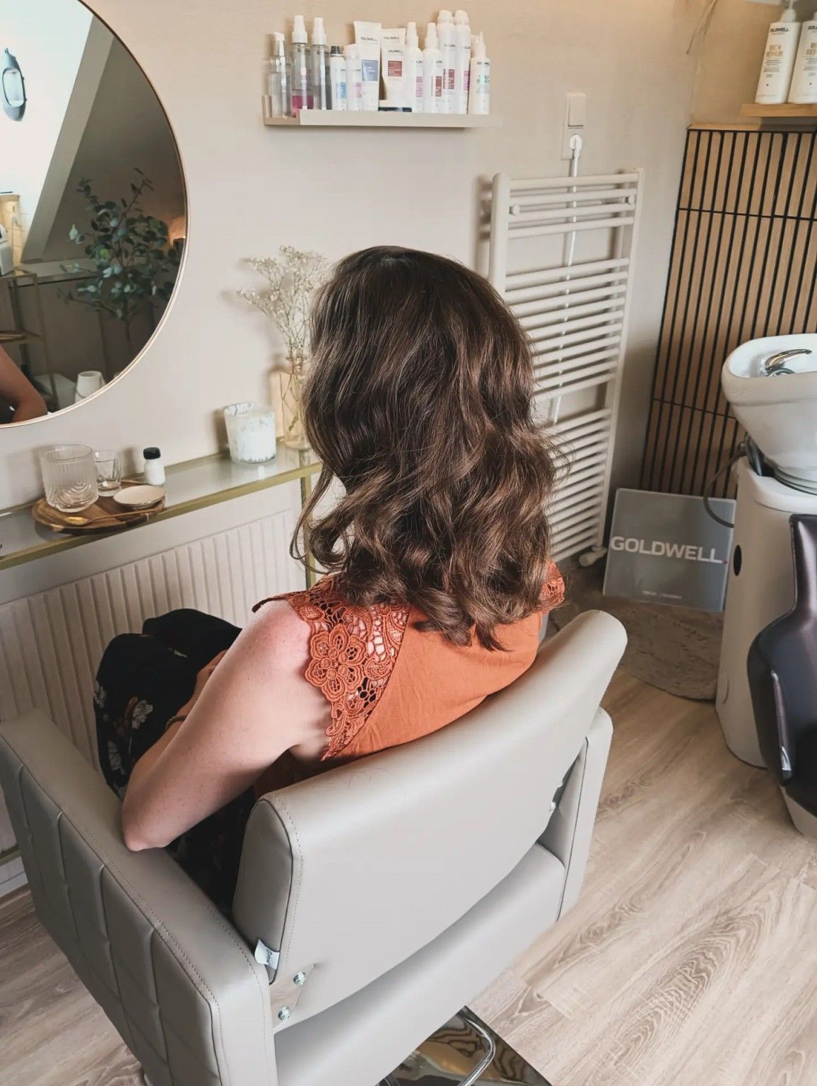 Persoon met golvend bruin haar zit in een lichtgrijze stoel in een lichte kapsalon, met het gezicht naar een spiegel.