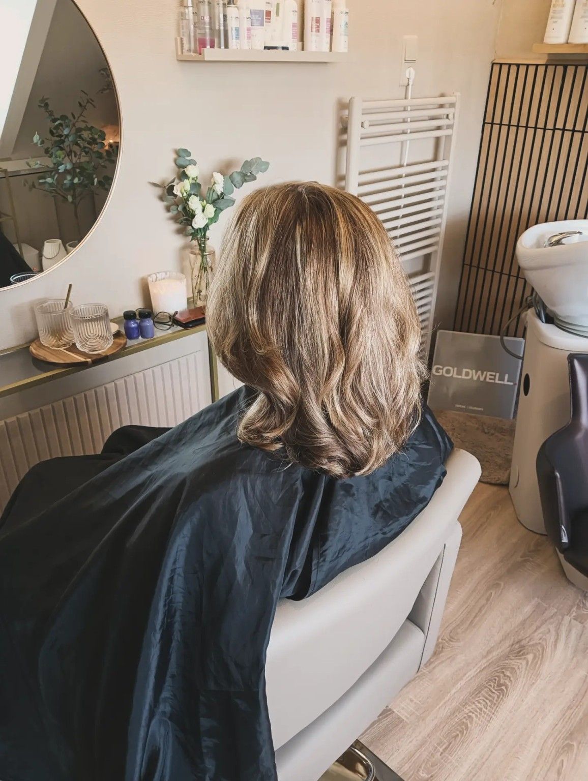 Persoon zit in een kappersstoel voor een spiegel, met een kappersmantel om en gestyled bruin haar.