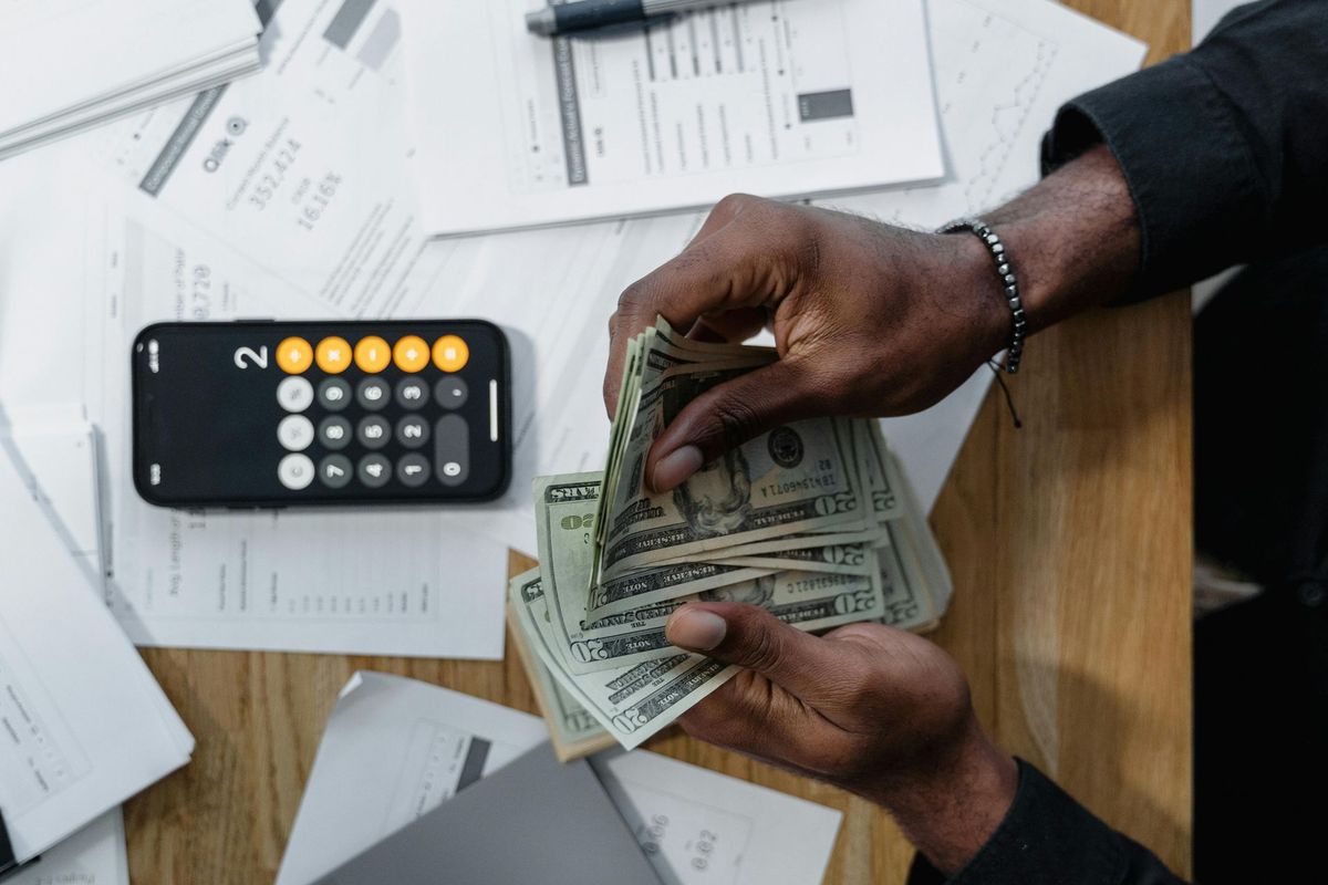 Hands counting a stack of US dollar bills next to a calculator and financial documents.