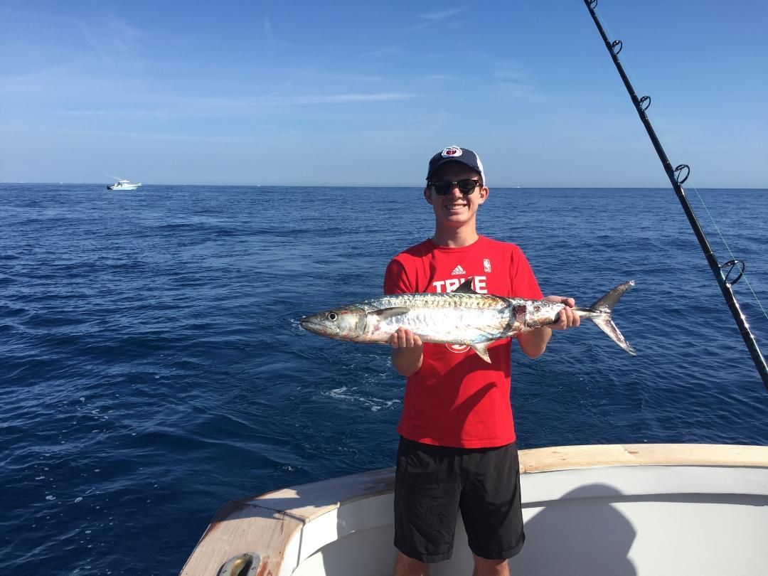 A person in a red shirt holding a freshly caught fish on a boat in the open ocean under a clear blue sky.