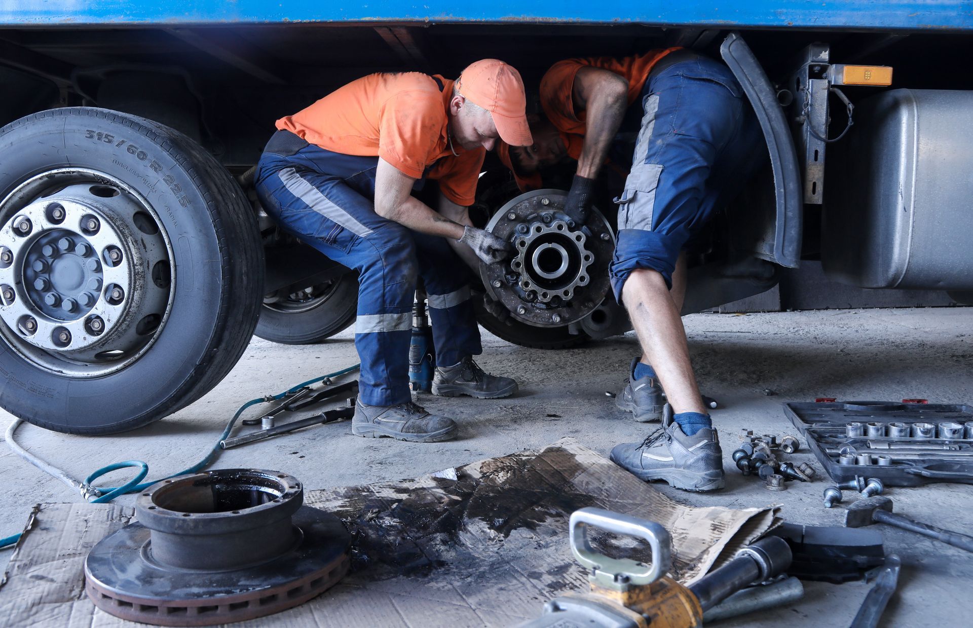 Two mechanics in orange shirts and blue pants repairing a large truck wheel assembly inside a garage