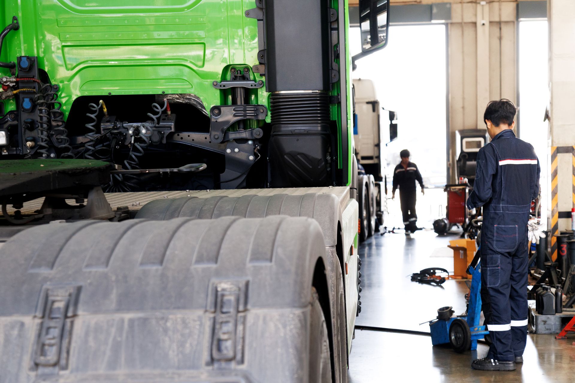 A mechanic wheeling a jack past a green semi tractor in a service bay for emergency trailer repair. A mechanic wheeling a jack past a green semi tractor in a service bay for emergency trailer repair.