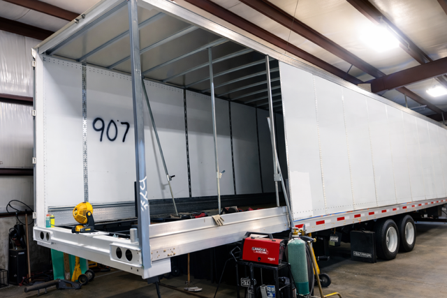 White box trailer with open rear door in a warehouse, numbered 907, showing its empty interior.