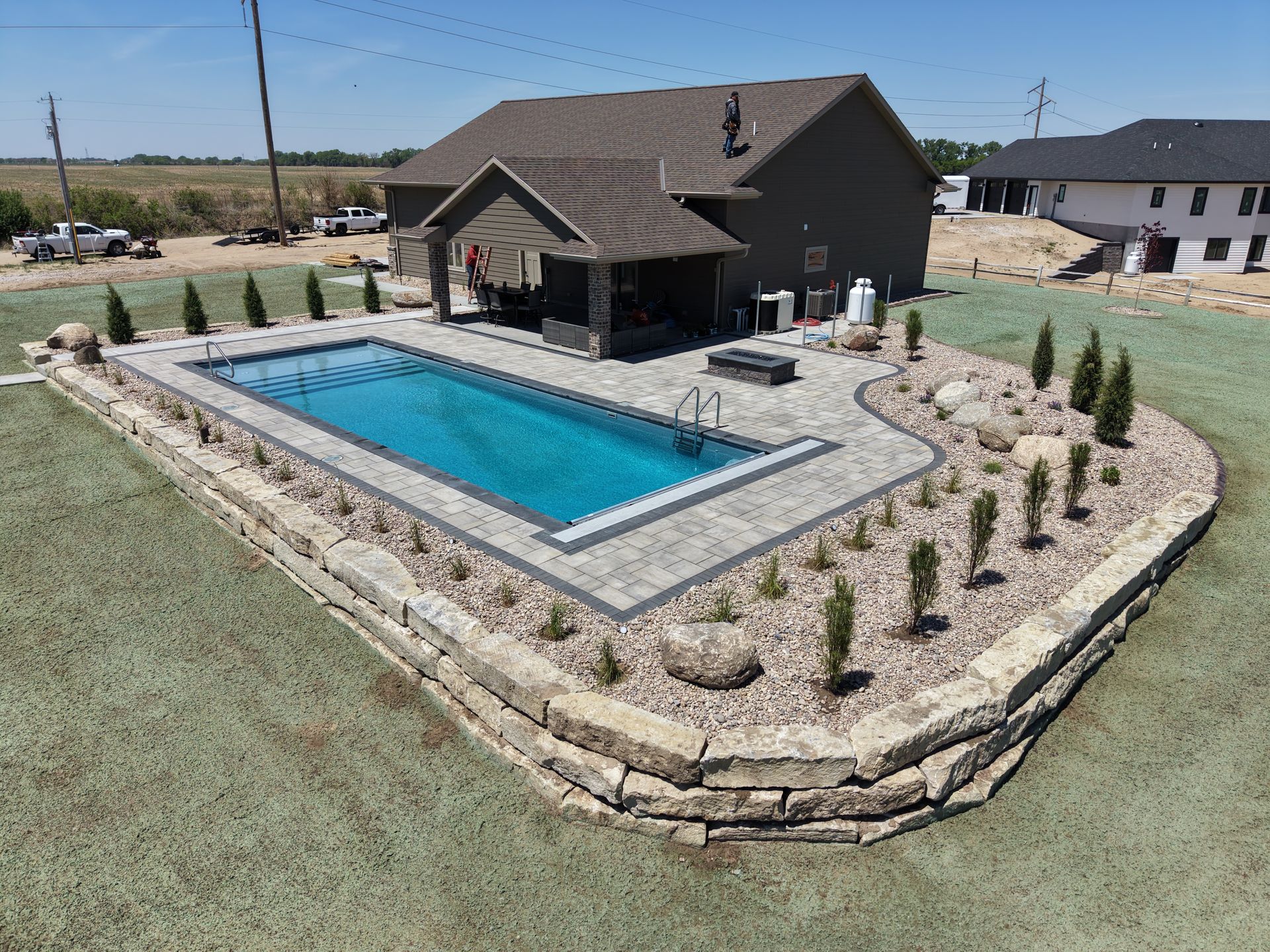 Backyard with rectangular blue pool, stone deck, and brown house on a landscaped lot.