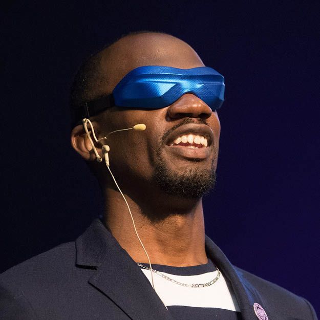 Lex Gillette wearing blue visor glasses and a headset microphone on stage, smiling in a dark suit.
