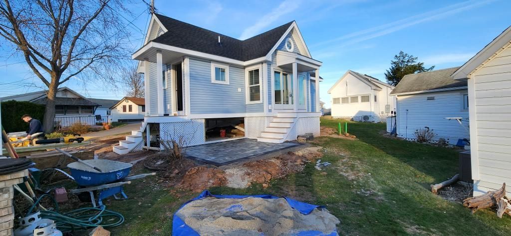 Blue-gray house under construction with a small yard, patio stones, and neighboring homes on a sunny day