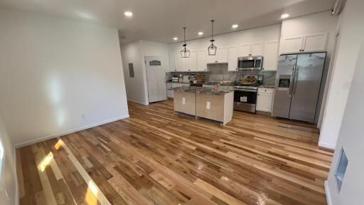 Open-concept kitchen and living room with hardwood floors, white walls, island, pendant lights, and stainless appliances