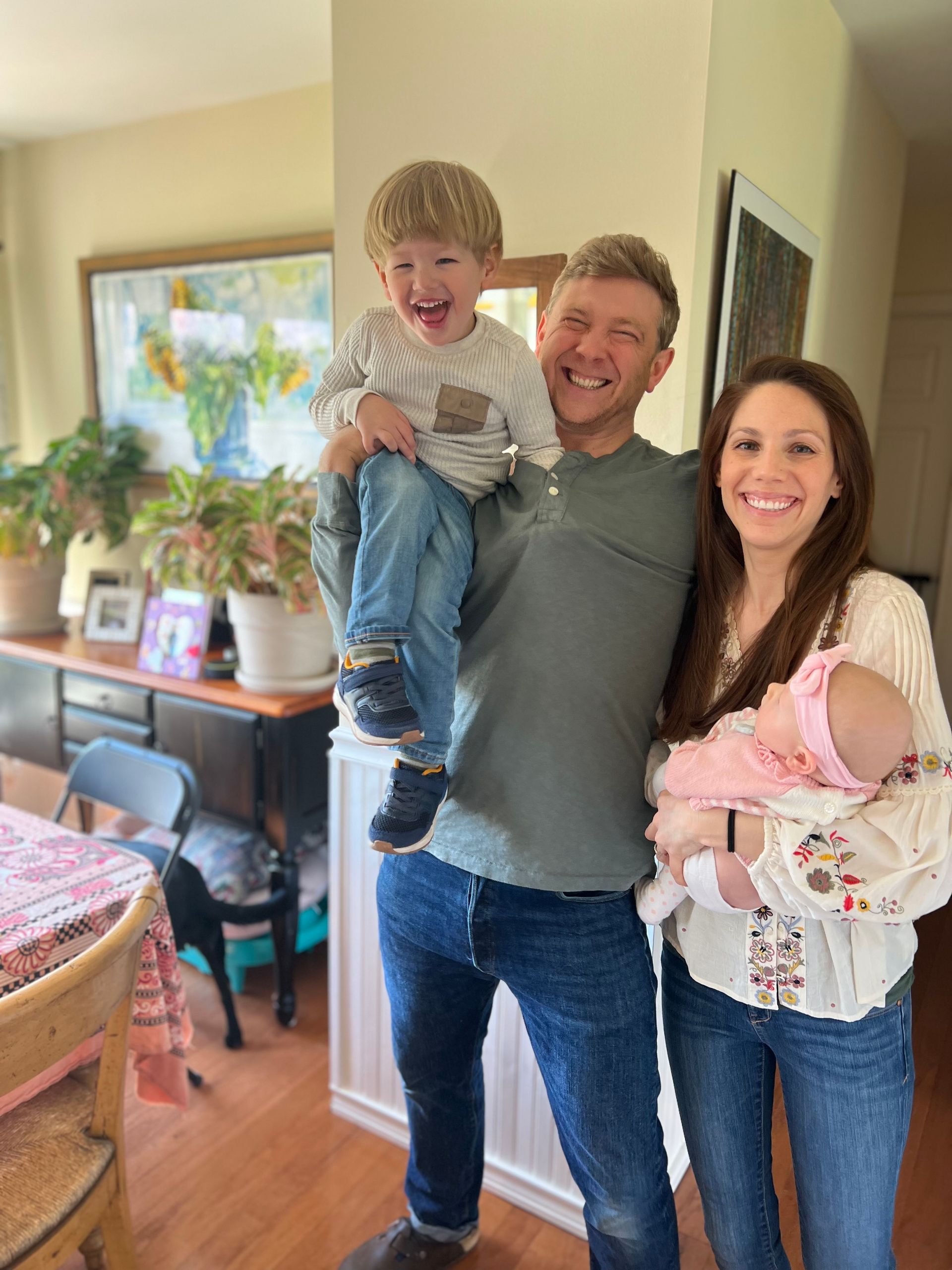 Family of four smiling indoors, holding a toddler and baby in a bright living room.