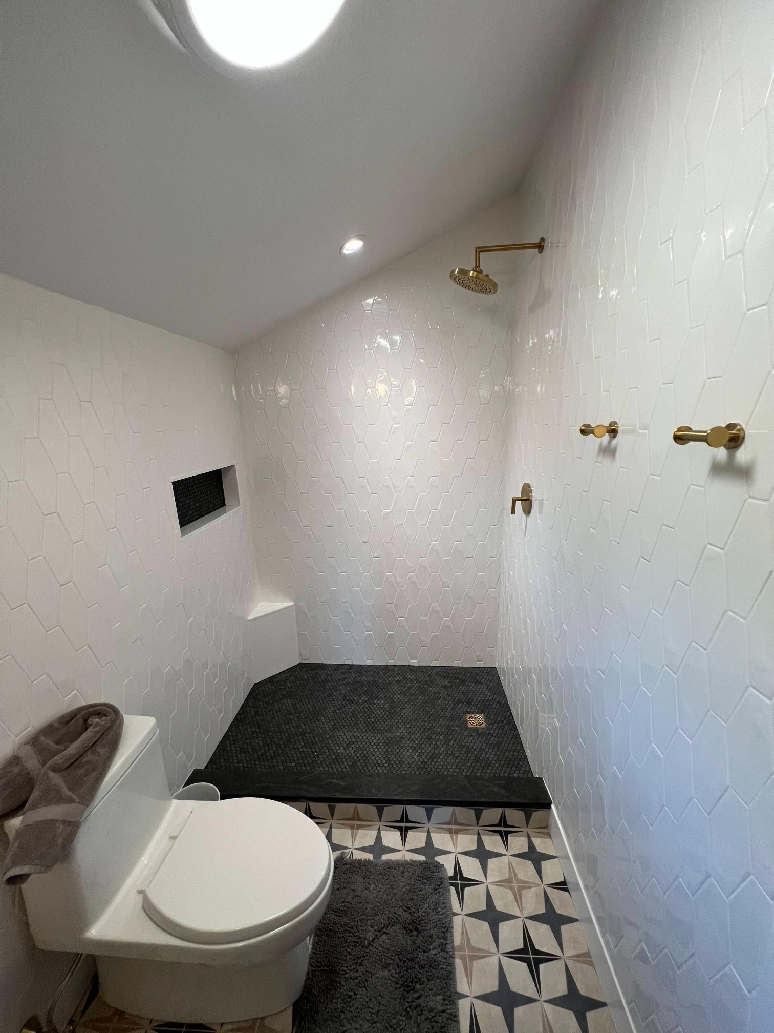 Small white bathroom with toilet, black rug, patterned floor, and shower at the back.