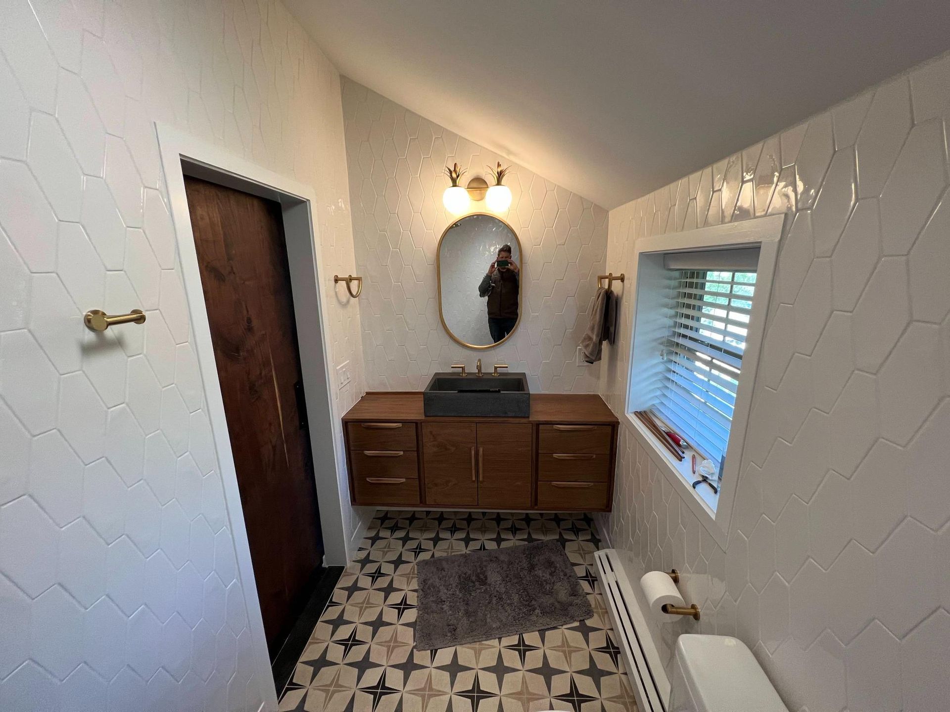 Small bathroom vanity with oval mirror, wood cabinet, patterned tile floor, and window with blinds.