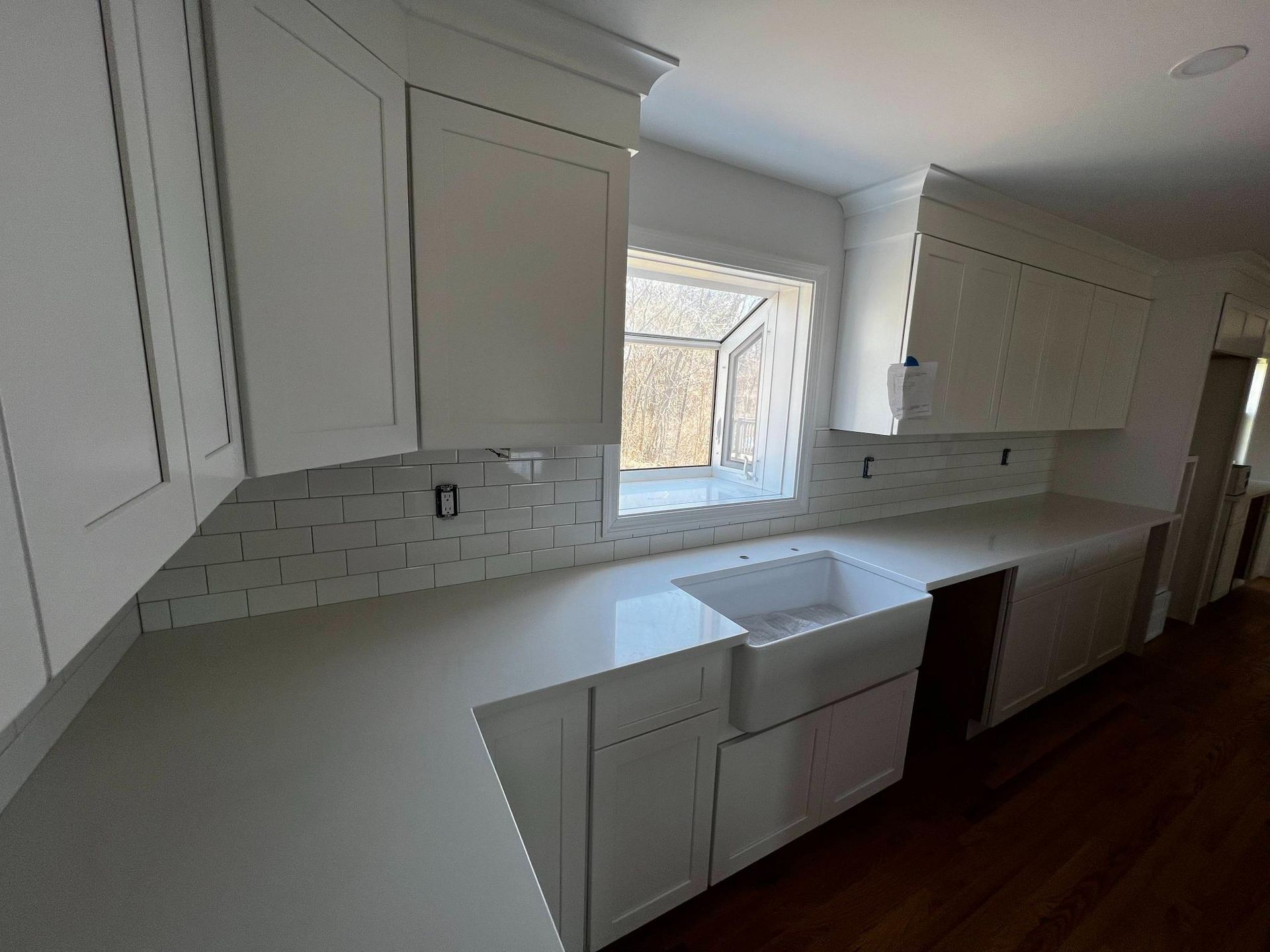 Bright white kitchen with cabinets, subway tile, sink, and window above a dark wood floor.