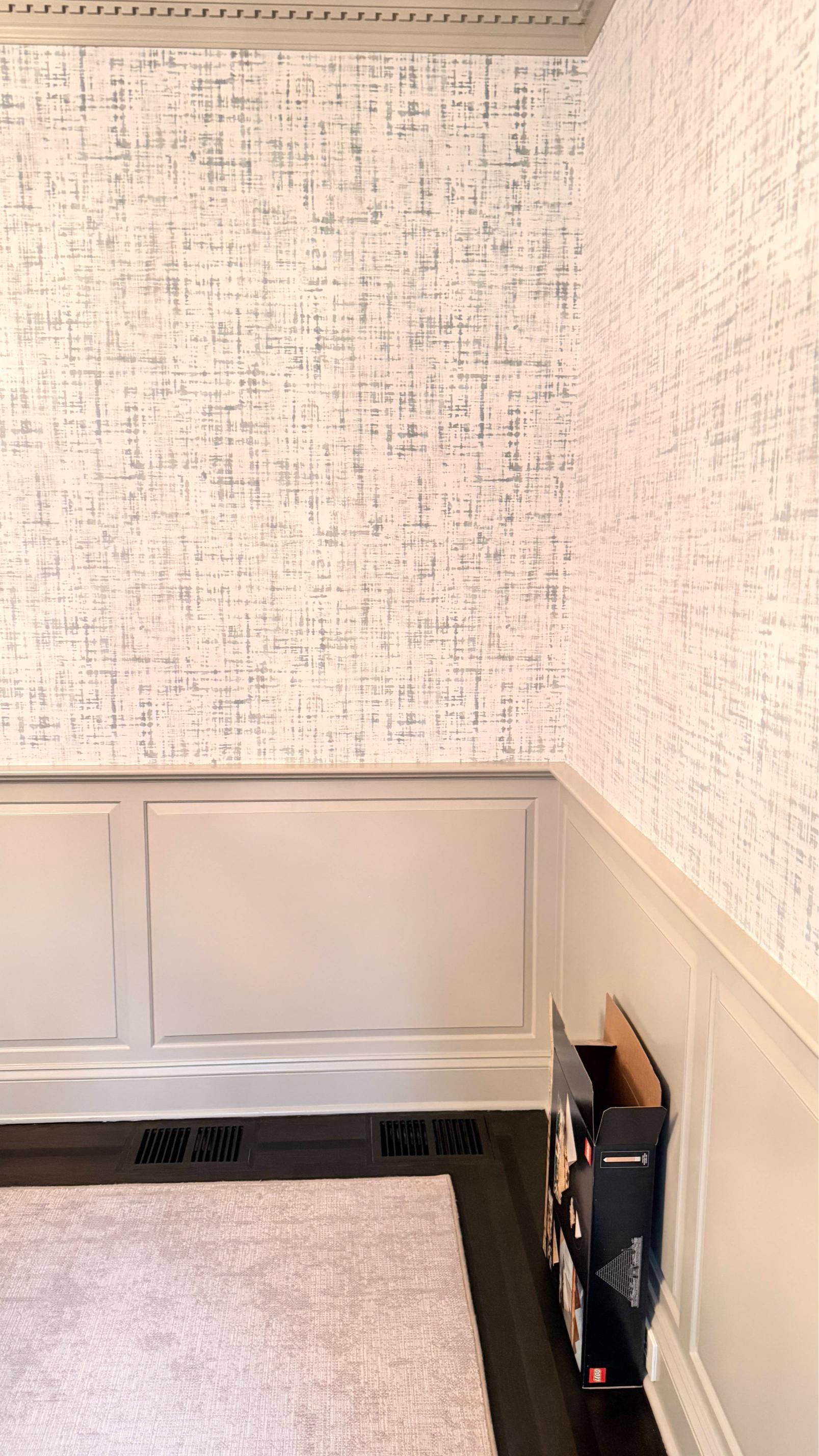 Empty corner of a room with patterned wallpaper, white wainscoting, gray floor, and a small wall-mounted device.