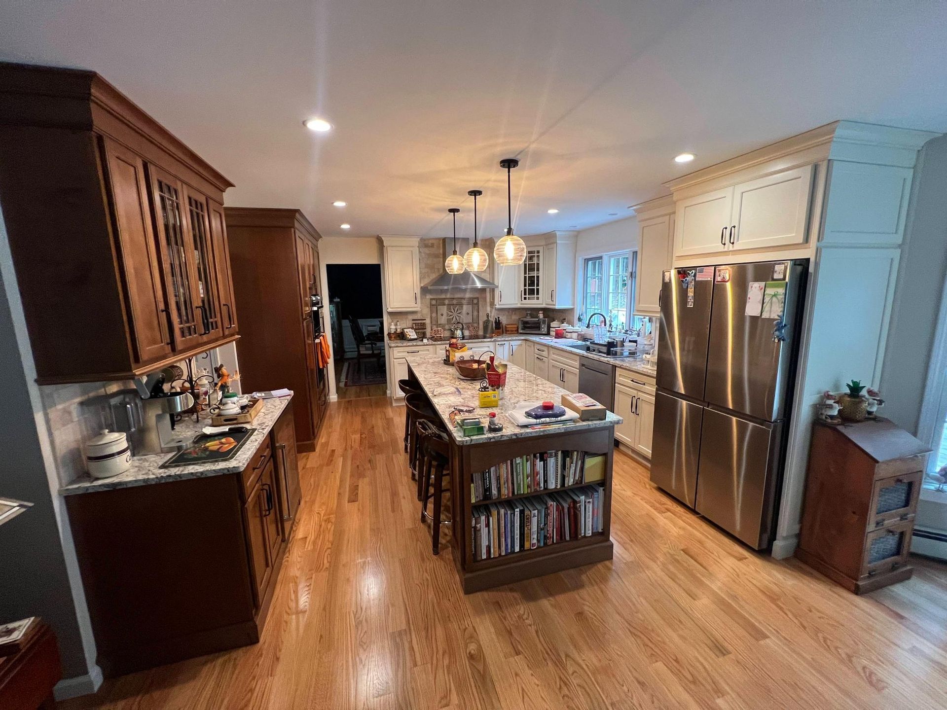 Bright kitchen with island, wood floors, stainless fridge, and dark cabinets; warm lights over a long room.