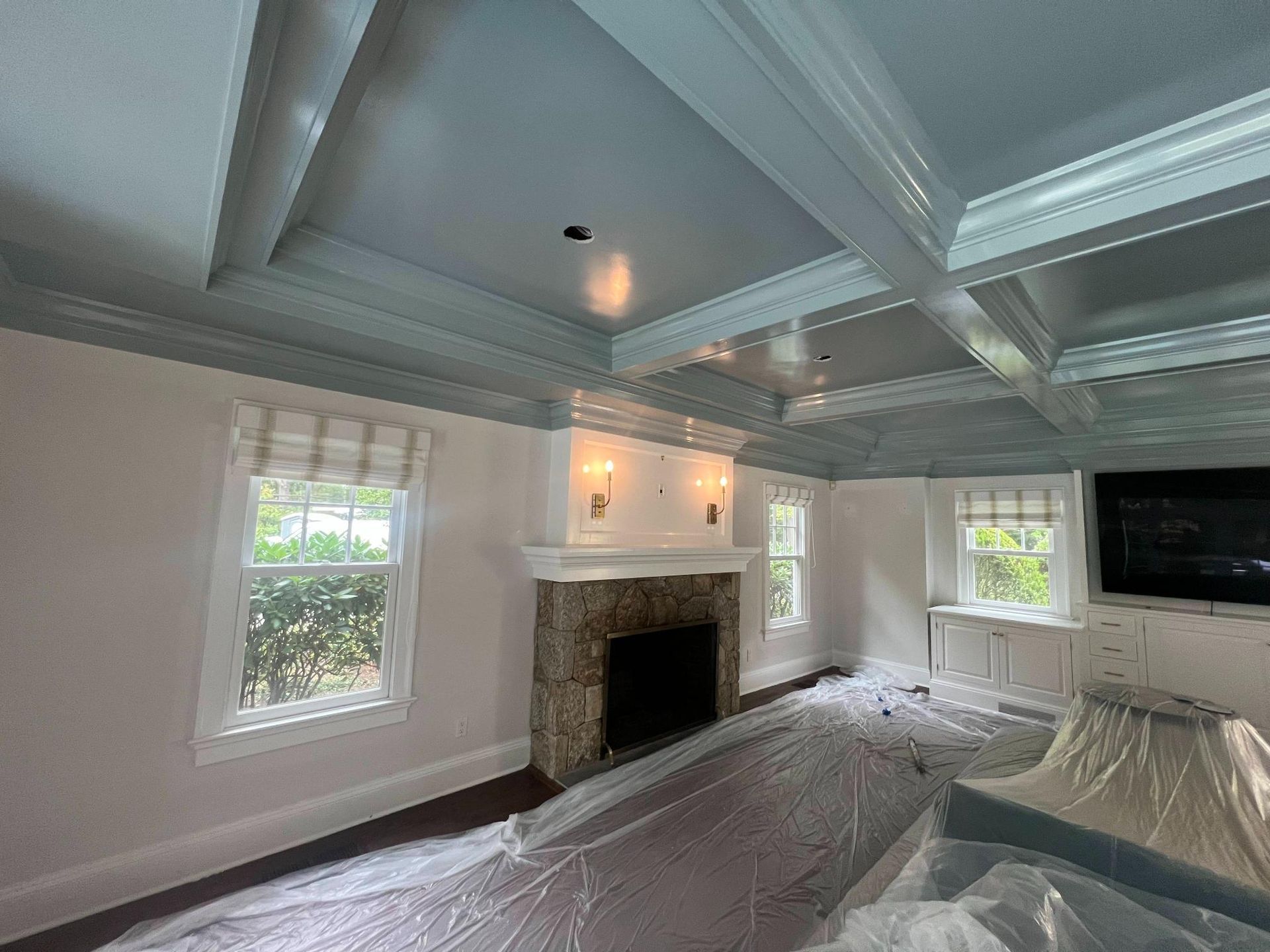 Empty living room with coffered ceiling, fireplace, windows, and plastic-covered floor under renovation