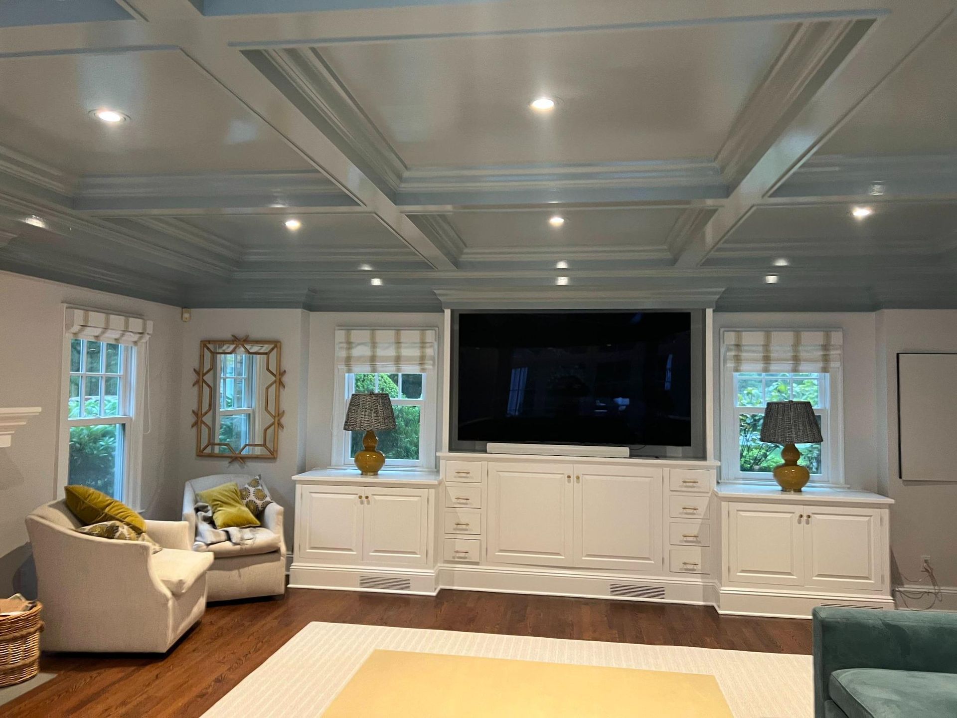 Bright living room with coffered ceiling, wall-mounted TV, white cabinets, and a gray armchair by windows