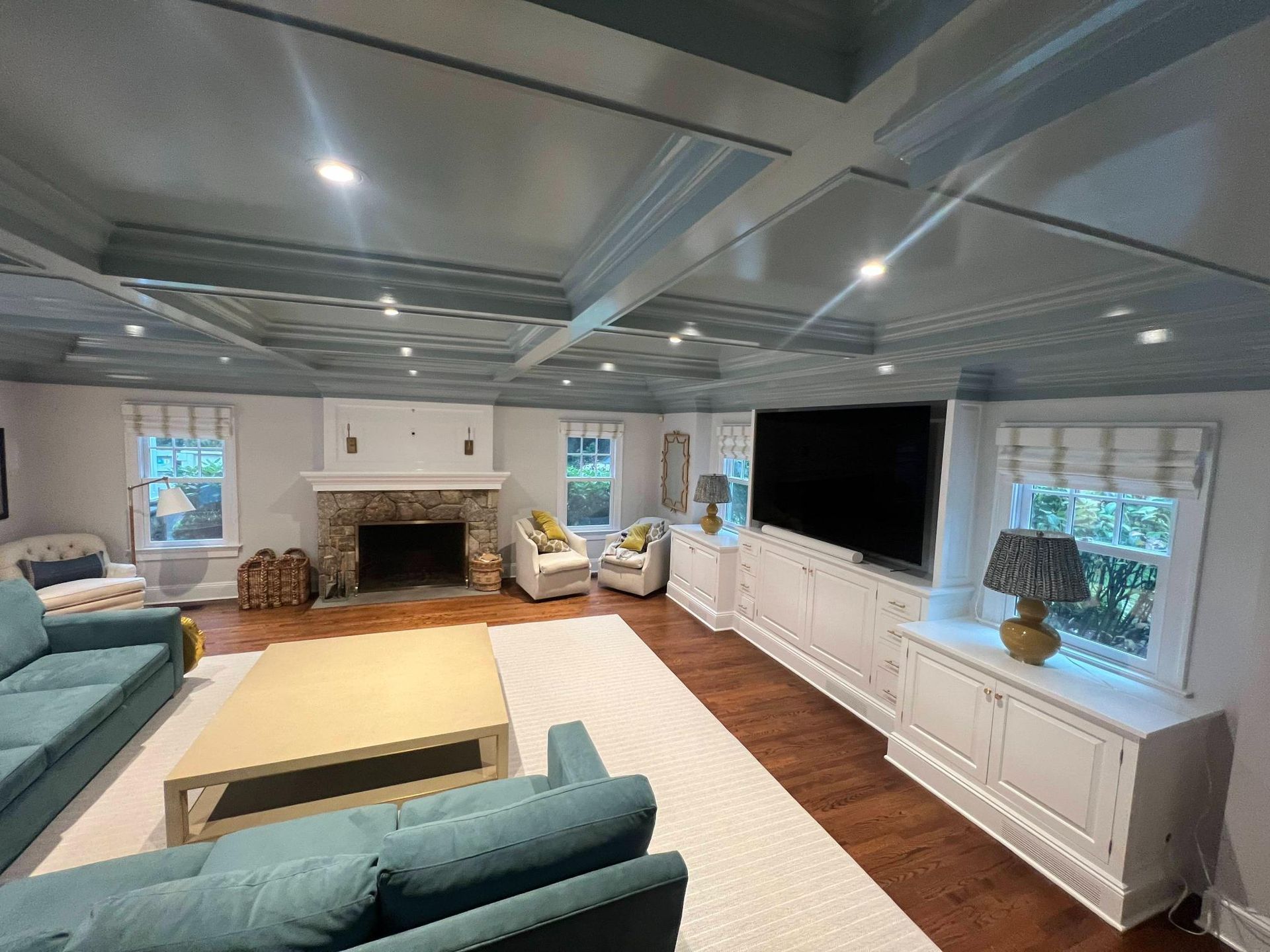 Bright living room with teal sofas, stone fireplace, large TV, white built-in cabinets, and recessed ceiling lights
