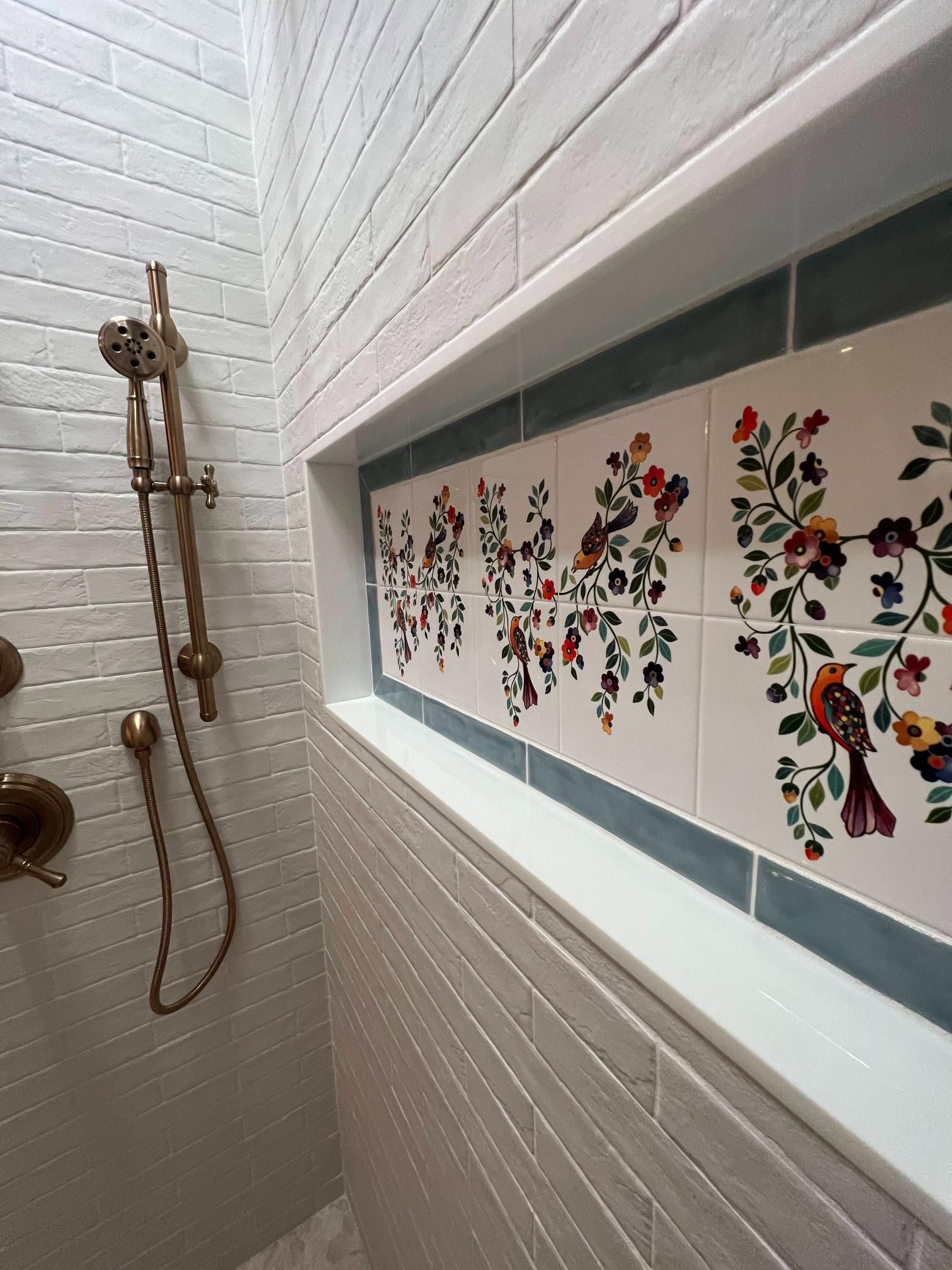 White shower with brass fixtures and a floral tile border in a narrow tiled bathroom.