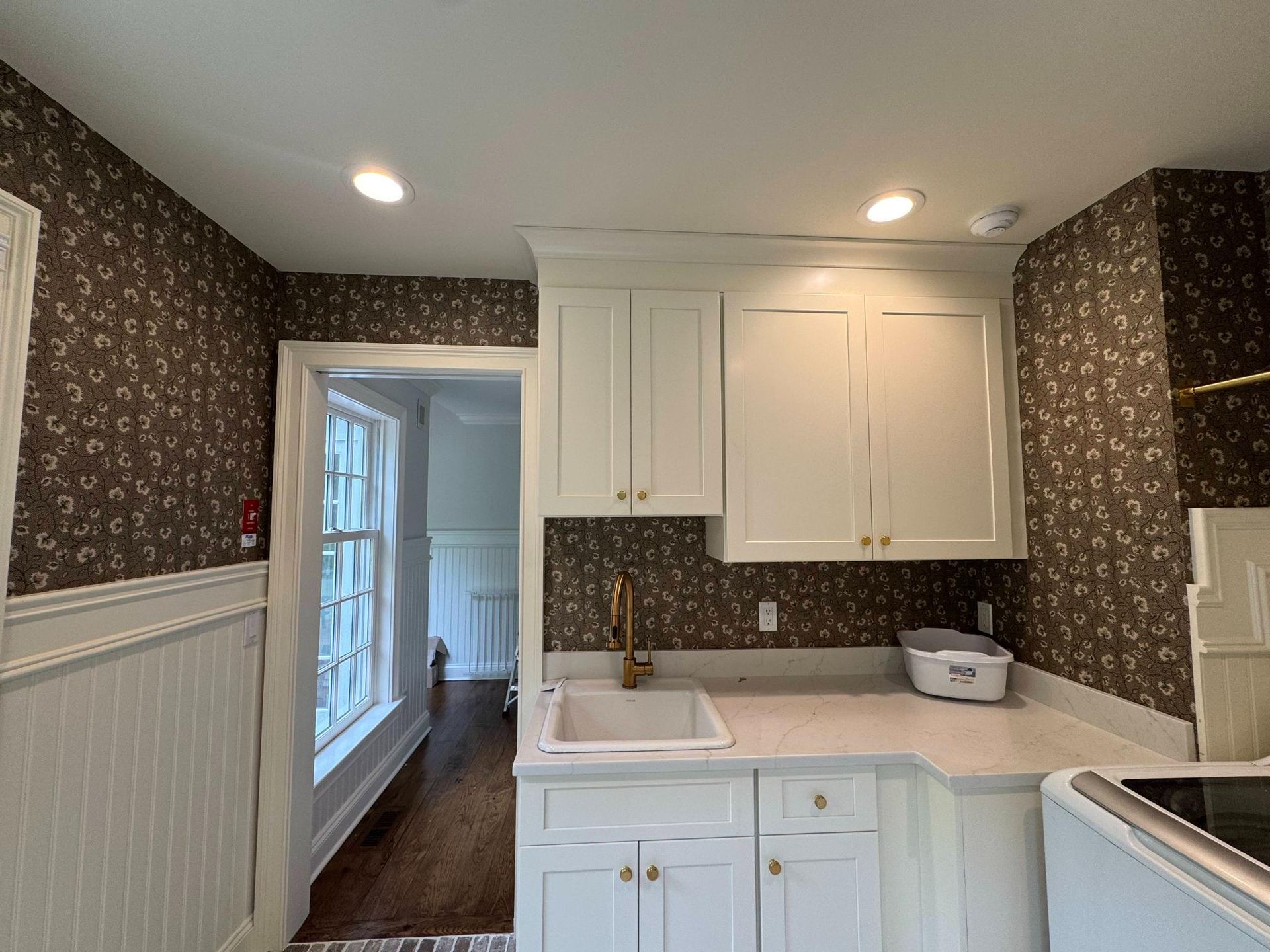 Small kitchen with white cabinets, dark patterned wallpaper, sink, and doorway to another room.
