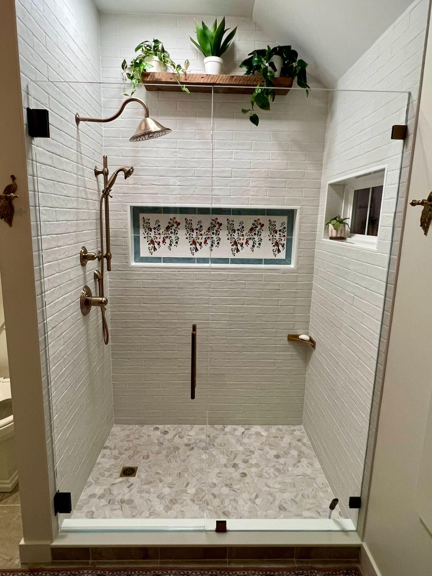Modern walk-in shower with white tile, brass fixtures, pebble floor, and plants on a shelf