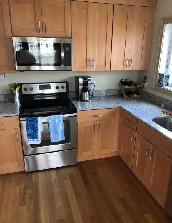 Modern kitchen with stainless steel appliances, wood cabinets, gray counters, and a window over the sink.