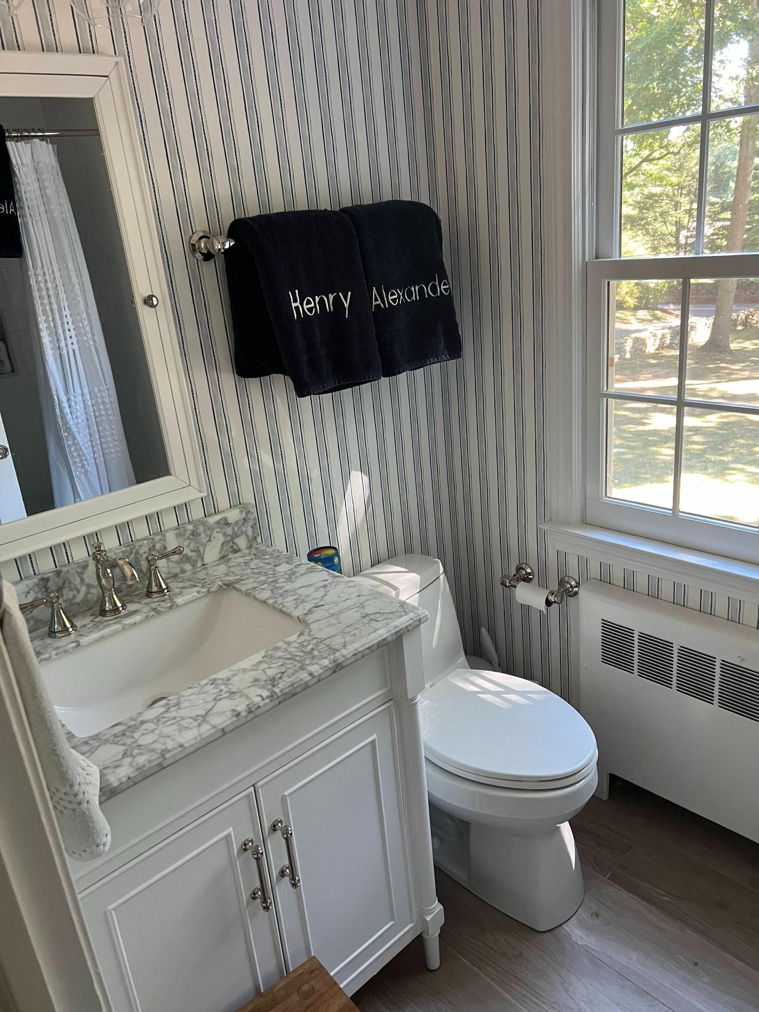 Small white bathroom with vanity, toilet, striped wallpaper, and towels by a window.