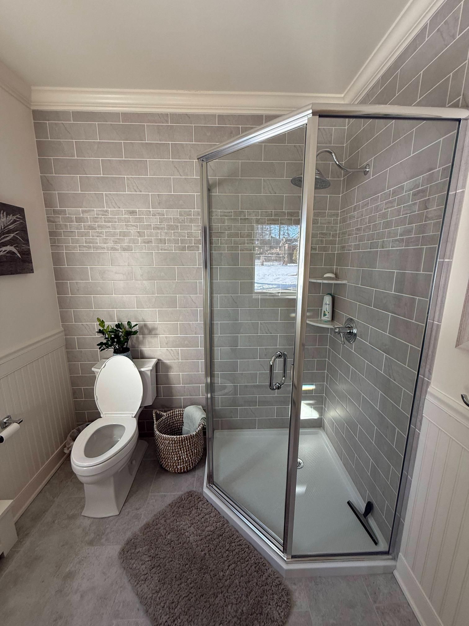 Small bathroom with glass corner shower, white tile walls, toilet, rug, and wicker basket.