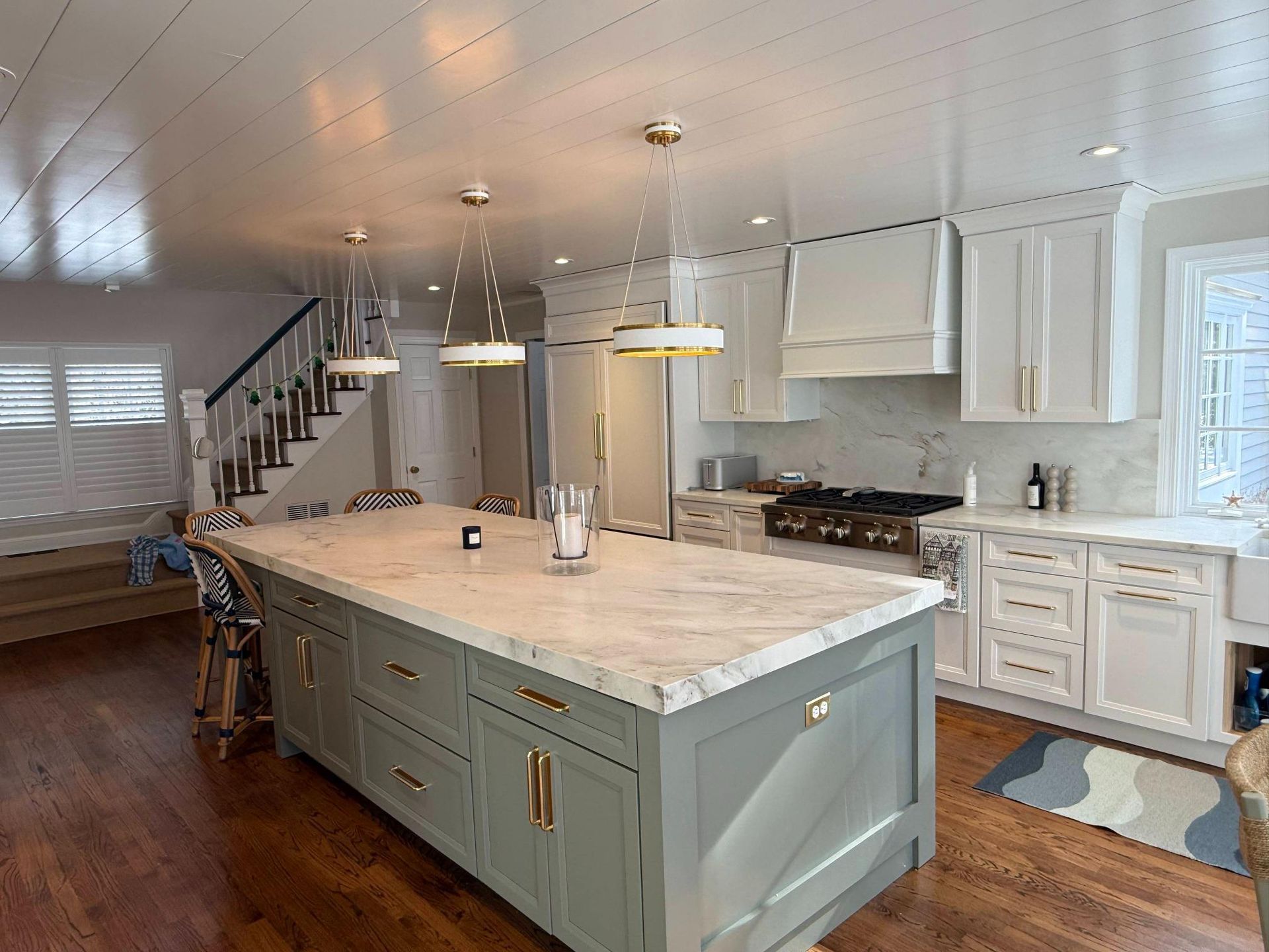 Bright kitchen with large island, white cabinets, hardwood floors, and stairs in the background