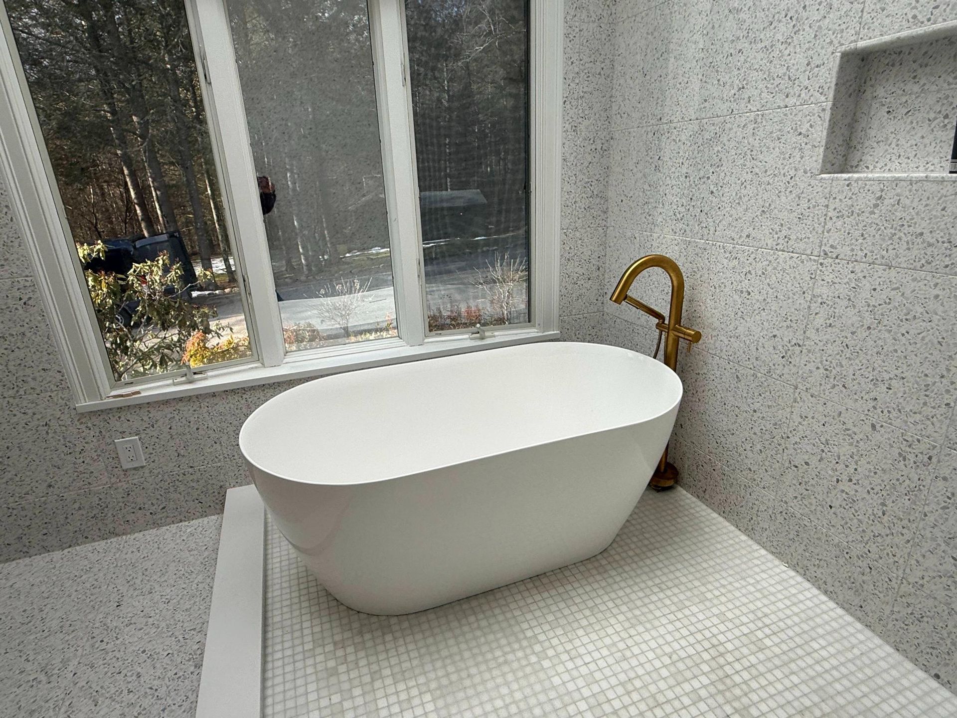 Modern bathroom with a white oval freestanding tub by a window and gold faucet