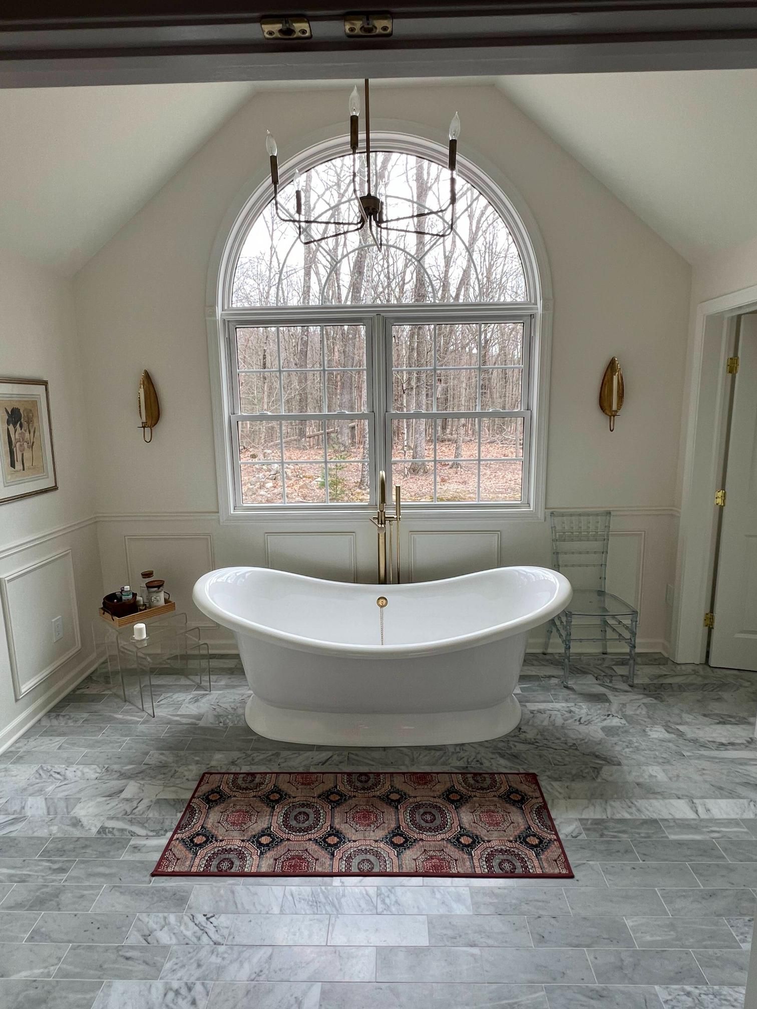 Bright bathroom with freestanding tub under arched window and patterned rug on marble floor