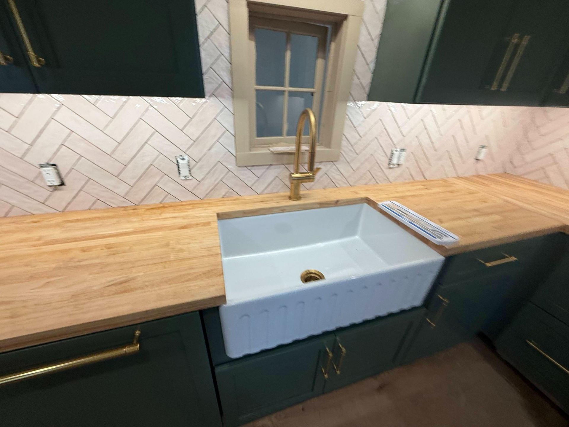 Modern kitchen sink with gold faucet, butcher-block countertop, and dark green cabinets under a window.