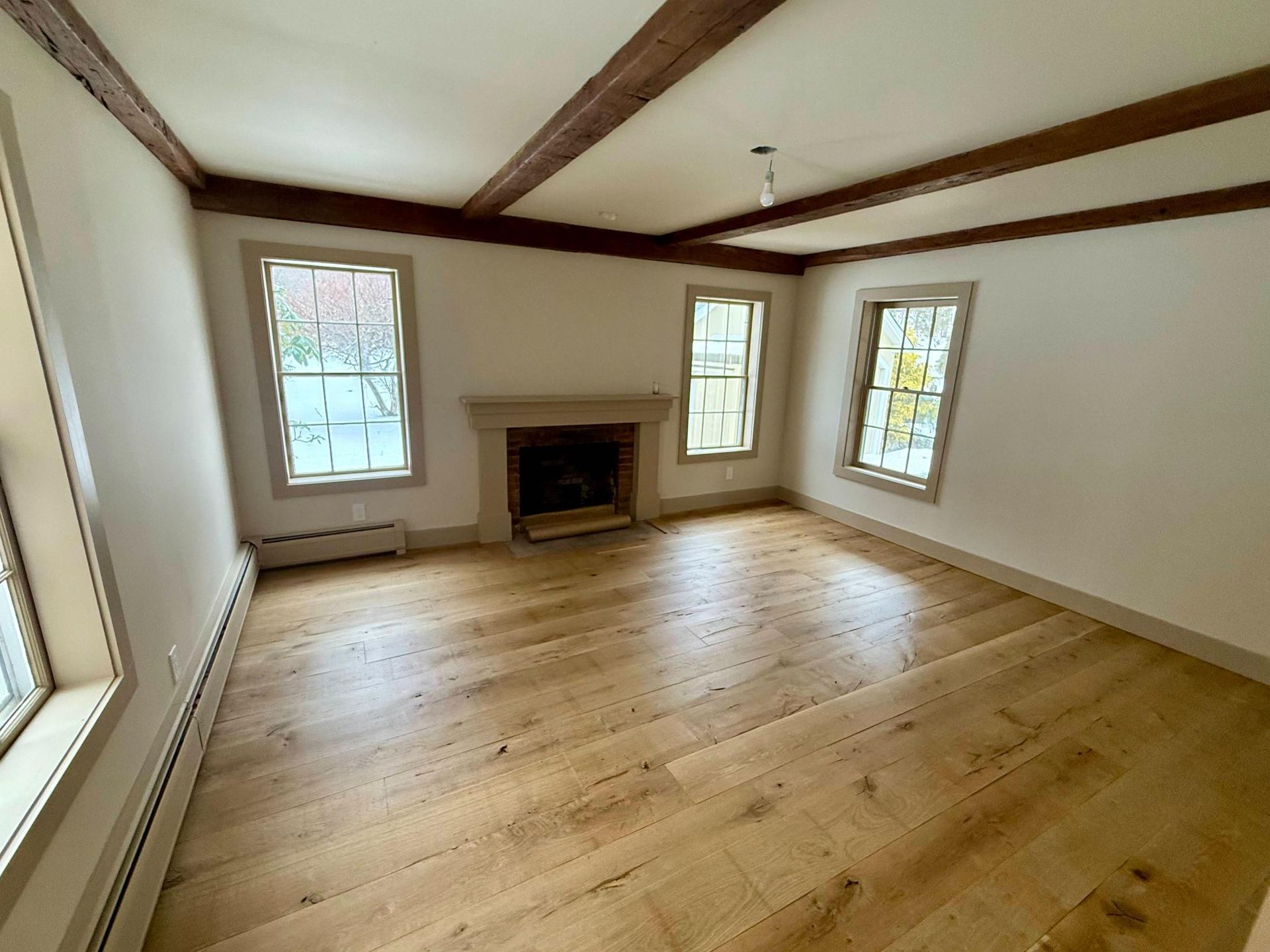 Empty living room with hardwood floors, white walls, exposed beams, and three windows around a fireplace.
