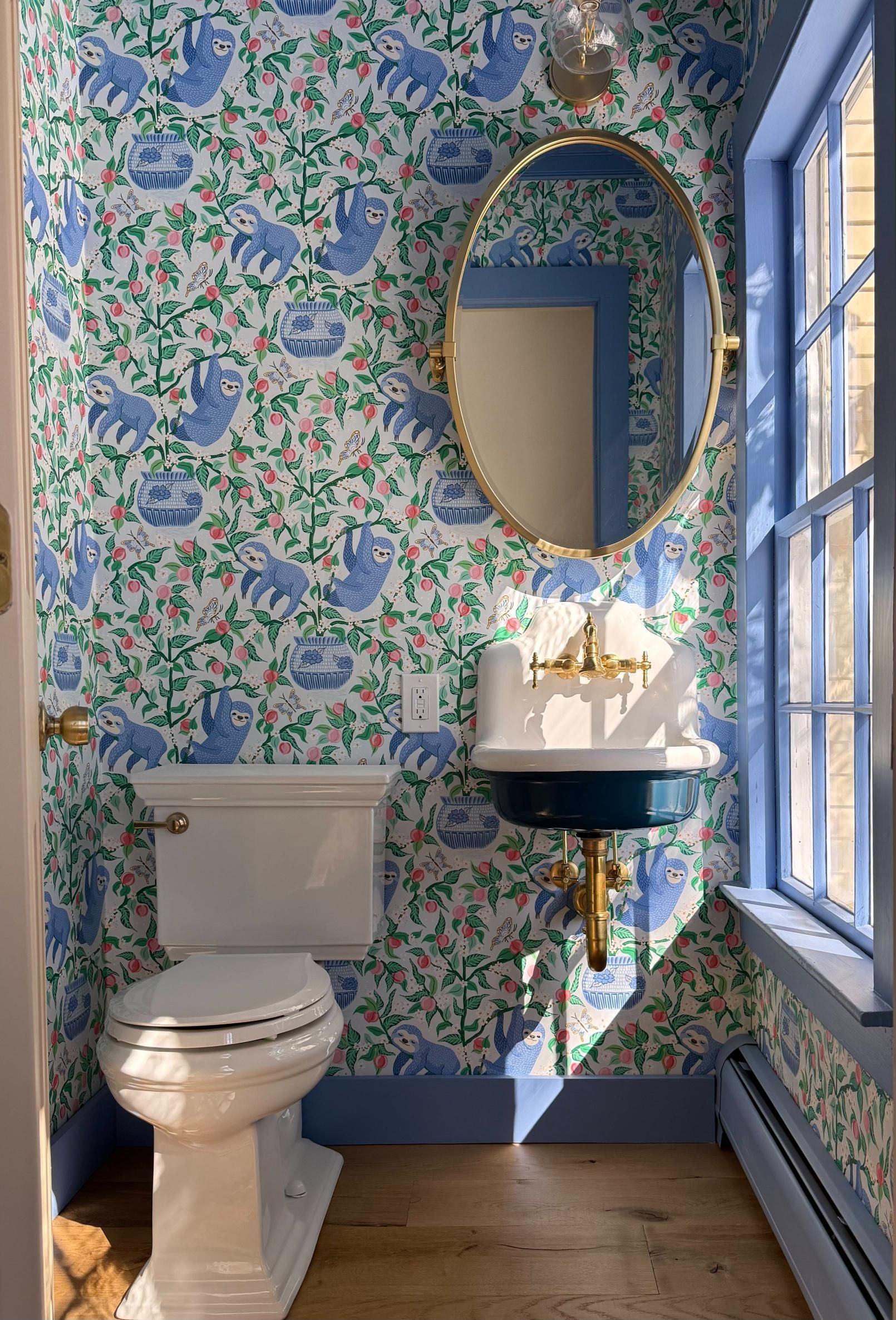 Small powder room with floral wallpaper, white toilet, oval mirror, pedestal sink, and blue-trimmed window.