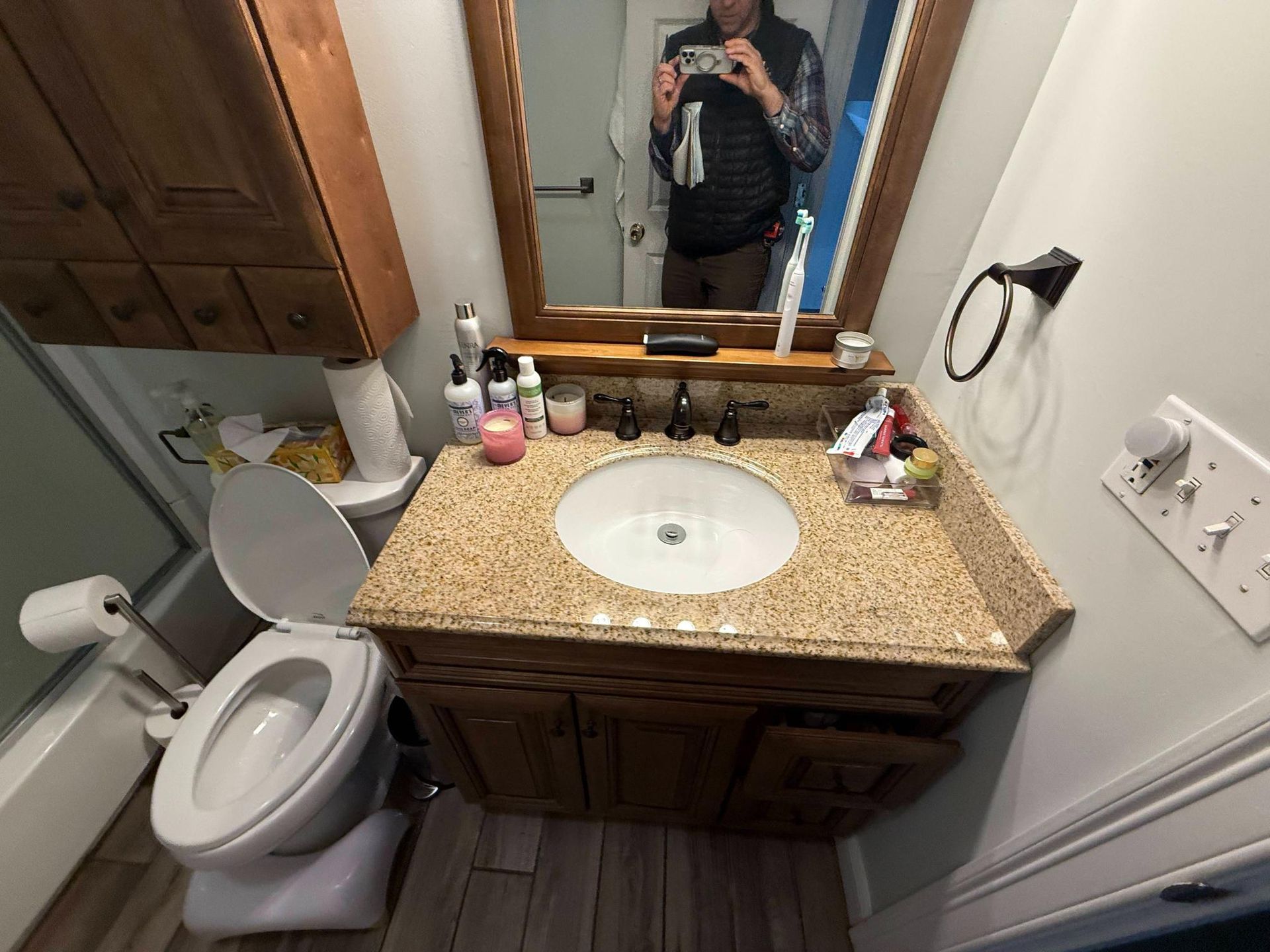 Small bathroom with granite sink vanity, toilet, mirror, and a person taking a mirror selfie.