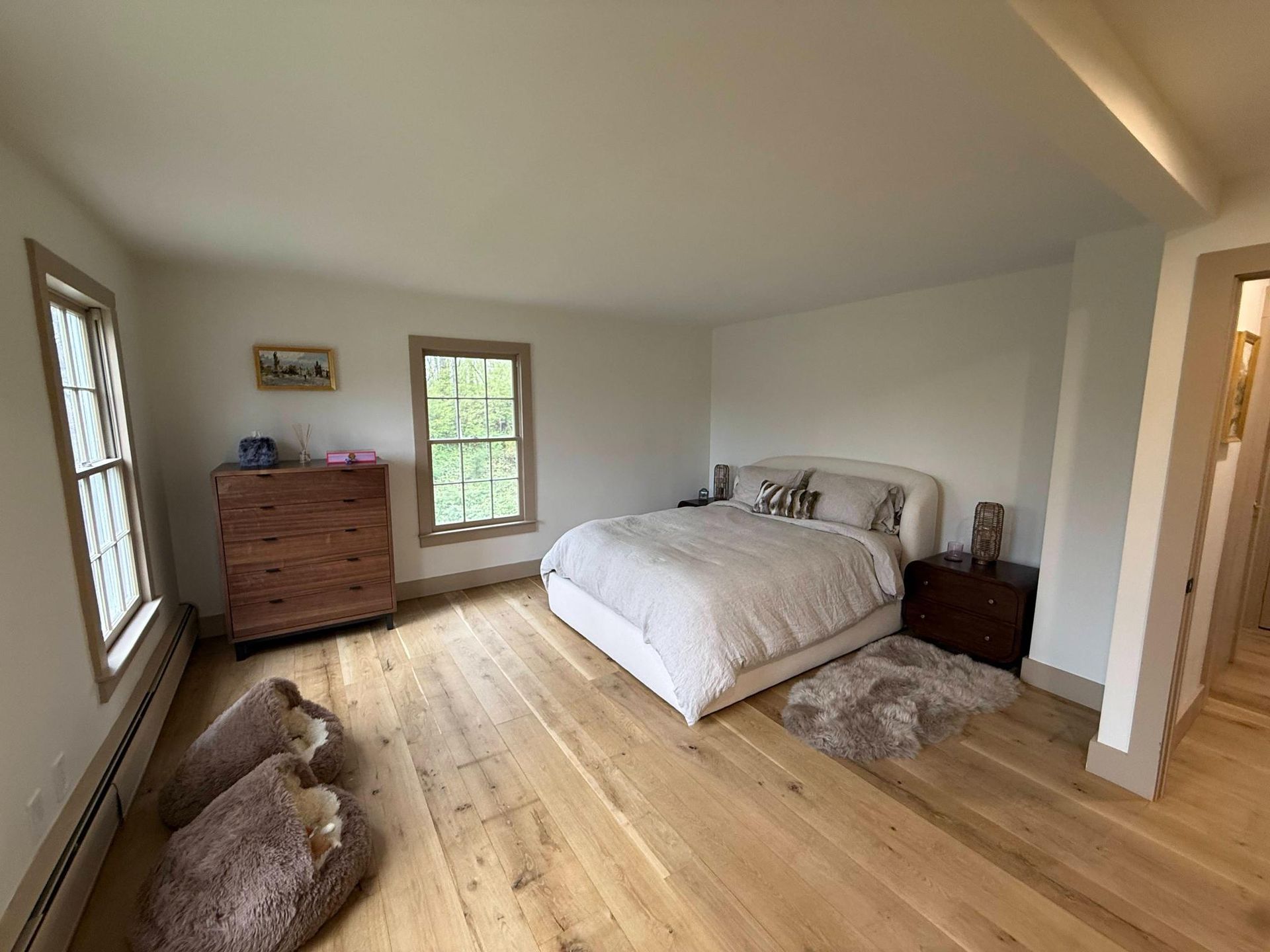 Spacious bedroom with white bed, wood dresser, large windows, and a wooden floor.