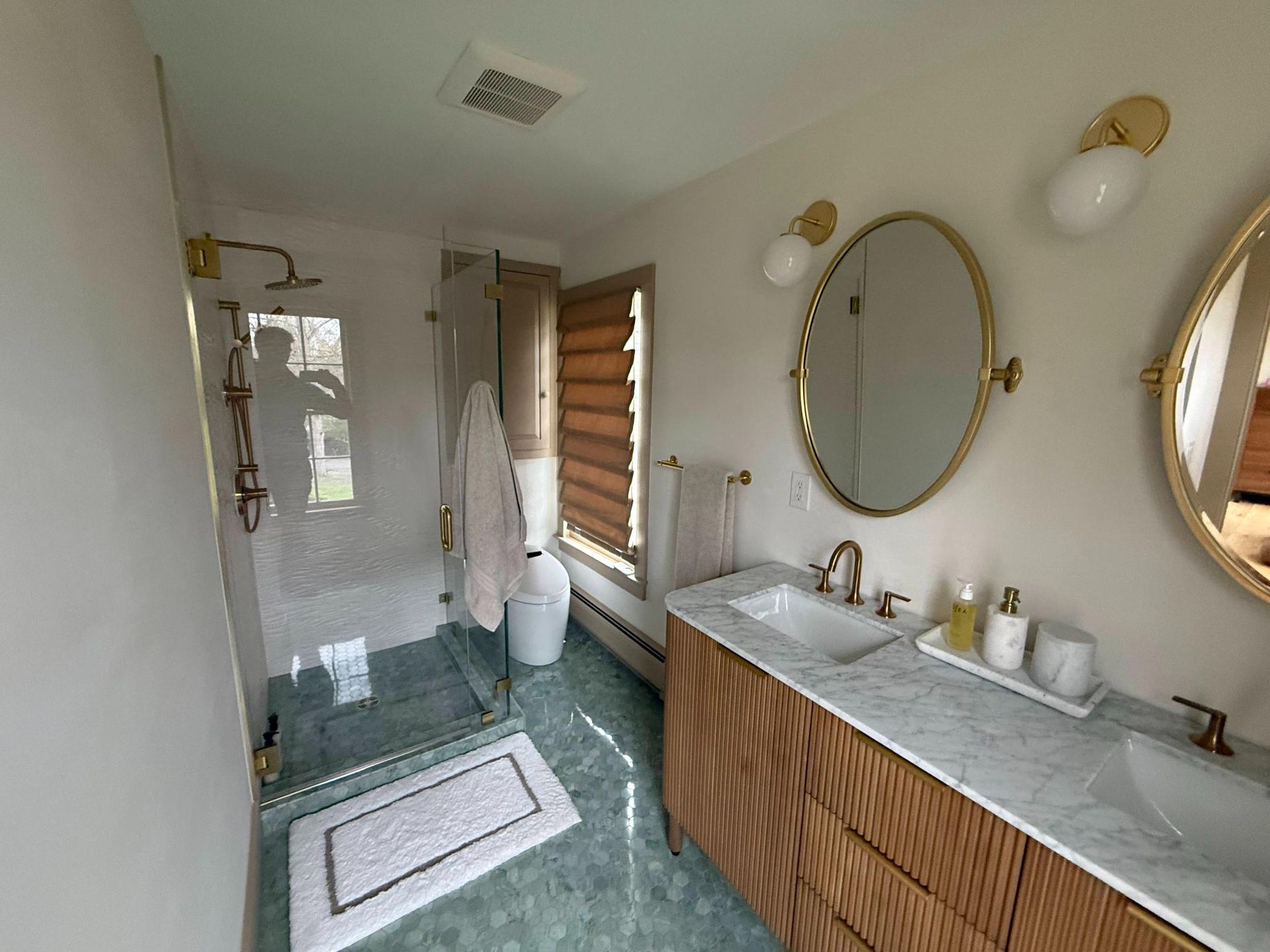 Bright bathroom with double gold mirrors, wood vanity, glass shower, and patterned rug