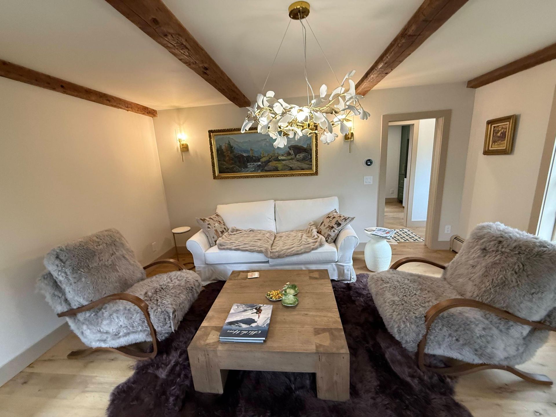 Cozy living room with sofa, two chairs, coffee table, chandelier, and wood-beamed ceiling.